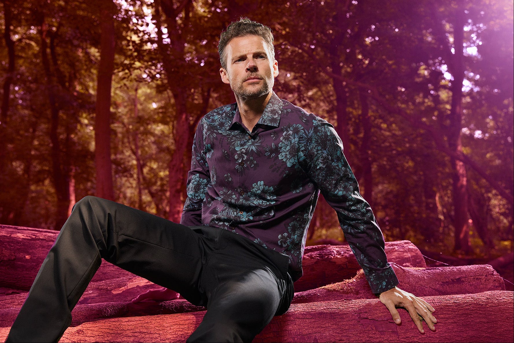 Man in a plum colored floral shirt sitting on a log in a forest with a purple hue