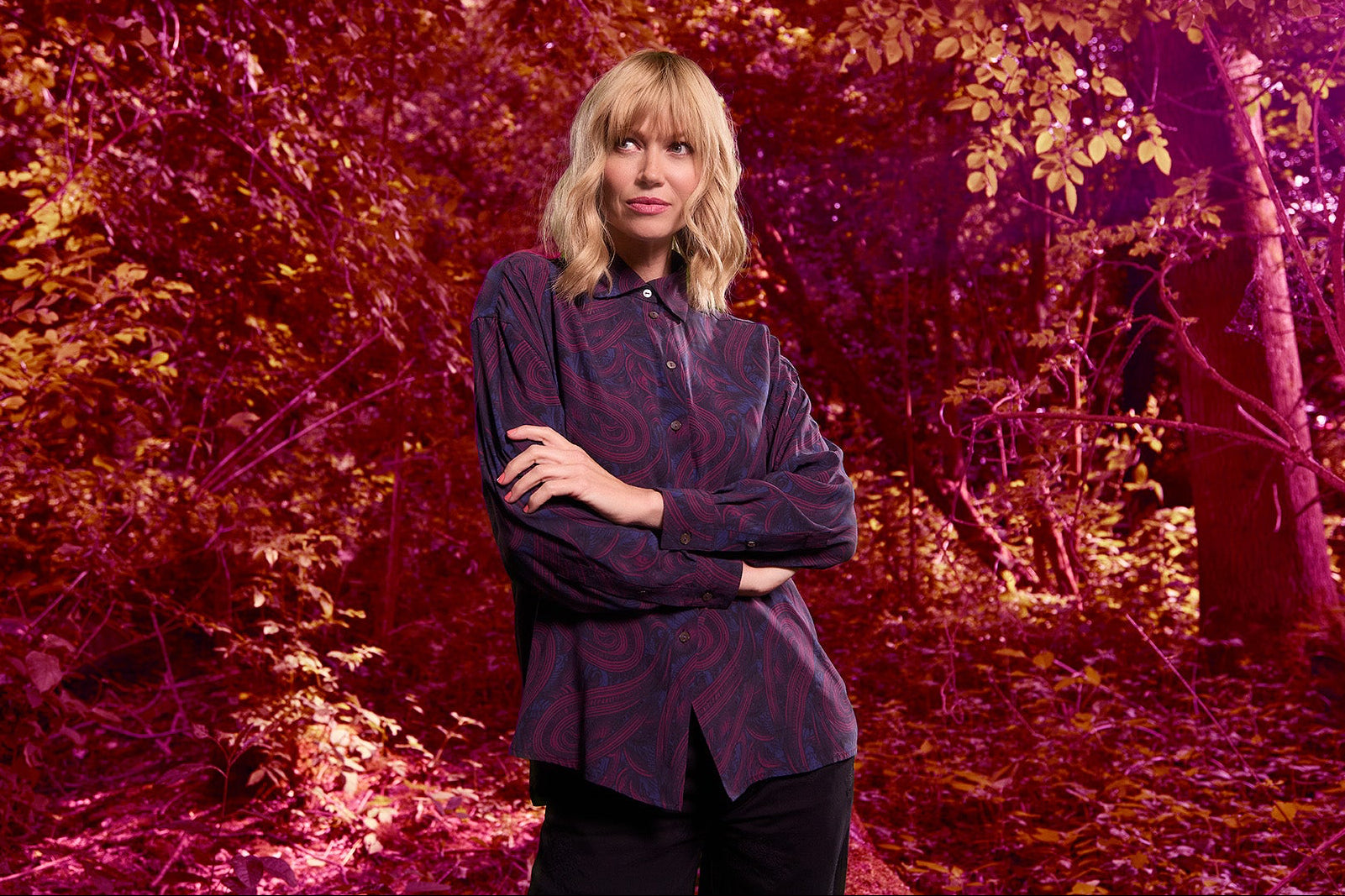 Woman in a plum paisley button down shirt standing in a forest with a red and purple color filter